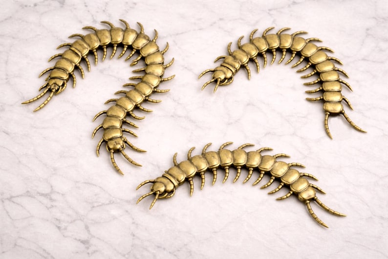 May include: Three antique gold-colored centipede decorative objects. Each centipede has a segmented body with numerous legs and antennae. The objects are arranged on a white and gray marble-patterned surface.