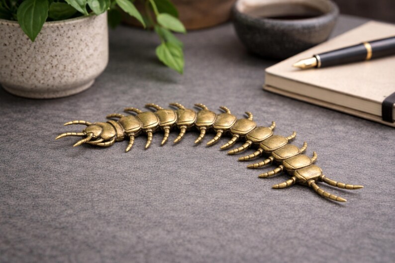 May include: A detailed, brass-colored centipede sculpture. The segmented body features multiple pairs of legs and antennae. The decorative piece is displayed on a gray surface, suggesting a desk or table setting.