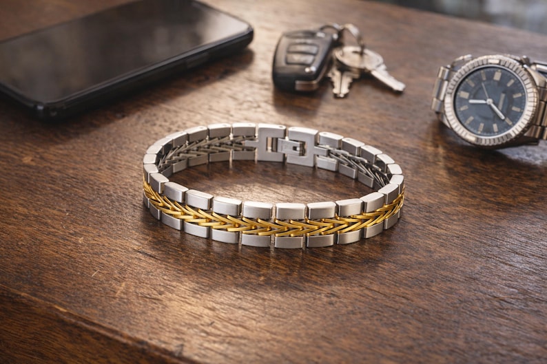 May include: A silver and gold bracelet with a woven gold centre section. The bracelet is on a wooden surface, with a watch, keys, and a smartphone in the background. The watch has a silver band and a dark face.