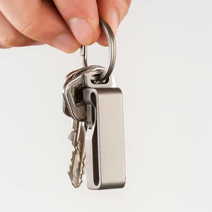 May include: A silver key organizer holding several keys. The organizer is rectangular with a curved design and a key ring. The keys are silver and have various shapes. The background is plain white.