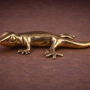 May include: A detailed, golden-colored metal lizard figurine. The lizard is posed on a brown surface, with its legs and tail extended. The figurine has a smooth, polished finish, and is likely a decorative item.