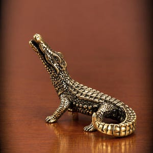 May include: A small, metallic, brass-colored figurine of an alligator in a standing position with its head tilted upward and mouth slightly open. The surface features detailed, textured scales across the body, limbs, and curved tail.