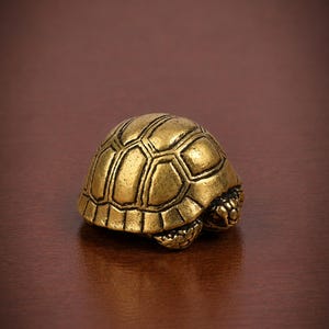 Op de afbeelding: Een klein, massief messing beeldje van een schildpad met een gedetailleerd, gestructureerd schild met geometrische schubben. Het metaal heeft een warme, gepolijste gouden afwerking met donkere accenten in de dieper gelegen delen.