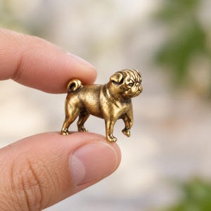 May include: A miniature brass pug figurine with a metallic gold finish, featuring a curled tail, wrinkled facial features, and a standing posture. The small sculpture is held between two fingers to demonstrate its compact size and scale.