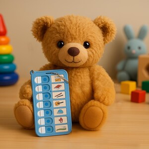 May include: A brown teddy bear holding a blue plastic communication board with various images and text. The board has a ring attached to it. The bear is sitting on a wooden surface with other toys in the background.