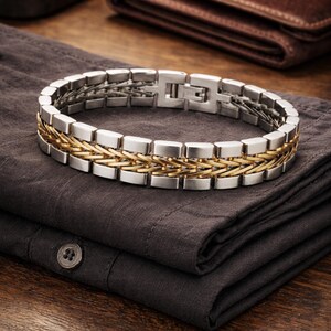 May include: A silver and gold-tone bracelet with a rectangular link design. The bracelet features a woven gold-tone centre section and a silver-tone clasp. The jewellery is displayed on a dark grey shirt, with a brown leather wallet in the background.