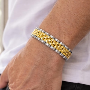 Könnte beinhalten: Ein zweifarbiges Armband mit Silber- und Golddesign. Das Armband besteht aus rechteckigen Gliedern mit abwechselnden Reihen aus Silber und Gold. Das Armband wird am Handgelenk getragen. Die Person trägt ein weißes Hemd und eine schwarze Jeans.