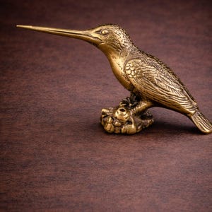 Op de afbeelding: Een gedetailleerde messing kolibrie sculptuur. De vogel zit op een getextureerde basis, met ingewikkelde veerdetaillering en een lange, slanke snavel. De sculptuur heeft een gouden tint en staat tegen een bruine achtergrond.