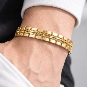 May include: A gold bracelet featuring a rectangular link design and a woven chain detail. The bracelet is worn on a wrist, with a white shirt and black garment visible in the background.