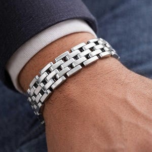 May include: A silver bracelet featuring a rectangular link design. The bracelet is worn on a wrist, with a dark blue blazer and white shirt visible in the background. The bracelet appears to be made of stainless steel.