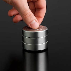 Könnte beinhalten: Ein silbernes Metall-Fidget-Spielzeug mit einem dreistufigen Design. Die oberste Ebene hat eine kreisförmige Vertiefung. Ein Finger drückt auf die Oberseite des Spielzeugs. Der Hintergrund ist schwarz.