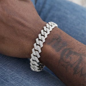 Puede incluir: Una pulsera plateada con un diseño de eslabones cubanos. La pulsera está adornada con pequeñas piedras brillantes. La pulsera se lleva en la muñeca sobre un fondo de mezclilla azul.