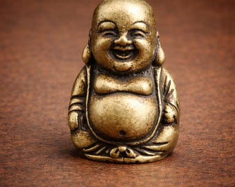 May include: A small, golden-colored Buddha statue in a seated position, smiling broadly. The figure has a rounded belly and is wearing a robe. The statue is likely made of metal and sits on a brown surface.