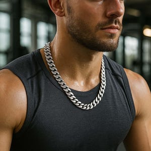 Puede incluir: Collar de cadena plateada con un diseño de eslabones gruesos. El collar se lleva sobre una camiseta sin mangas negra. La imagen está tomada en un gimnasio, con equipos borrosos en el fondo.