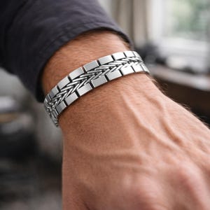May include: A silver bracelet with a woven design and rectangular links. The bracelet is worn on a person's wrist, with a dark grey shirt in the background. The bracelet is a piece of jewellery.