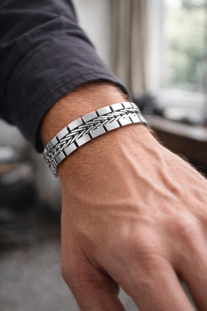 May include: A silver bracelet with a woven design and rectangular links. The bracelet is worn on a person's wrist, with a dark grey shirt in the background. The bracelet is a piece of jewellery.