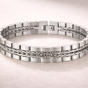 May include: A silver bracelet with a geometric design. The bracelet features rectangular links connected by a woven pattern. The clasp is a simple, secure design. The jewellery is displayed on a light background.