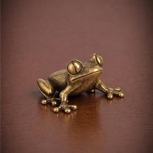 May include: A small, solid brass frog figurine with a textured, metallic finish. The frog is depicted in a seated, alert posture with large, prominent eyes, a wide mouth, and detailed webbed feet splayed outward on the ground surface.