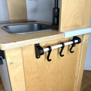 May include: A wooden play kitchen with a white sink, a black stove, and a white towel rack with black hooks.