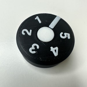 May include: A circular black control knob featuring a white center circle and white raised numbers one through five arranged around the perimeter. A white indicator bar is positioned between the numbers one and five on the textured surface.