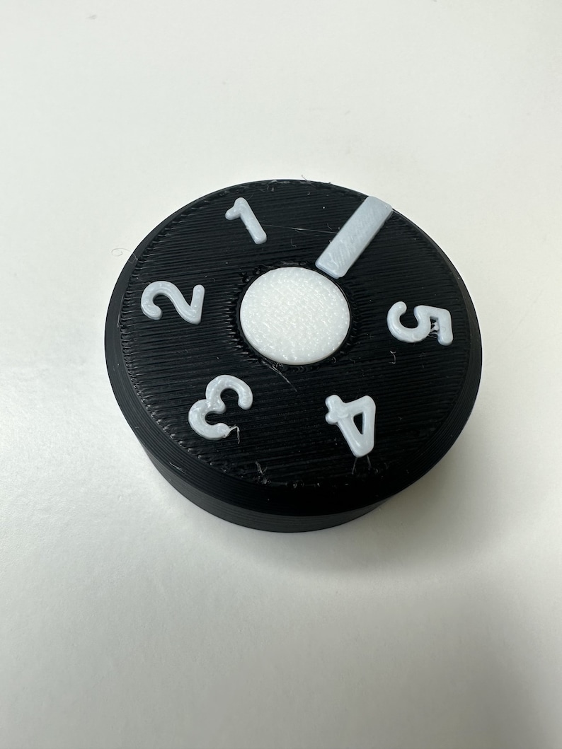 May include: A circular black control knob featuring a white center circle and white raised numbers one through five arranged around the perimeter. A white indicator bar is positioned between the numbers one and five on the textured surface.