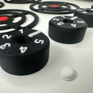 May include: Black cylindrical control knobs with white embossed numbers one through five and a white indicator line. The knobs feature a central circular opening and are arranged alongside black circular frames with red centers and small white discs.