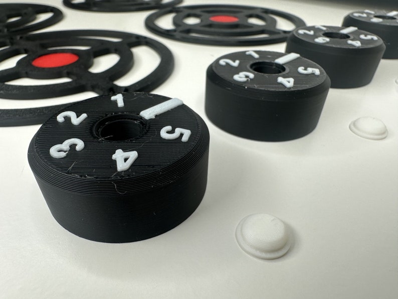 May include: Black cylindrical control knobs with white embossed numbers one through five and a white indicator line. The knobs feature a central circular opening and are arranged alongside black circular frames with red centers and small white discs.