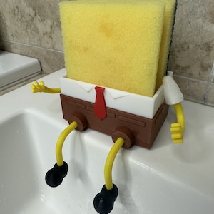 May include: A yellow sponge with a white shirt and red tie, sitting on a brown base with yellow legs and black feet. The sponge is in a sink.