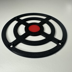 May include: A circular black plastic grill cover featuring a textured finish and a solid red center disc. The design includes four mounting holes positioned along the outer ring, with a cross-shaped support structure connecting the center to the frame.
