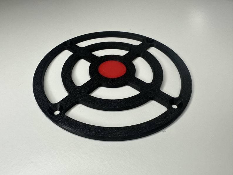 May include: A circular black plastic grill cover featuring a textured finish and a solid red center disc. The design includes four mounting holes positioned along the outer ring, with a cross-shaped support structure connecting the center to the frame.