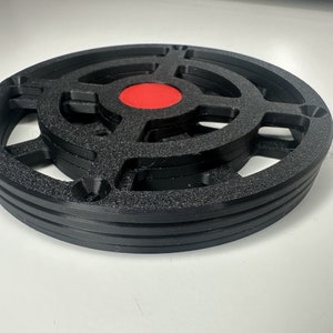 May include: A circular black 3D printed object with a textured surface, featuring a cross-shaped internal structure and a solid red circular center. The object has a layered design with visible horizontal ridges along the outer edge and four holes.
