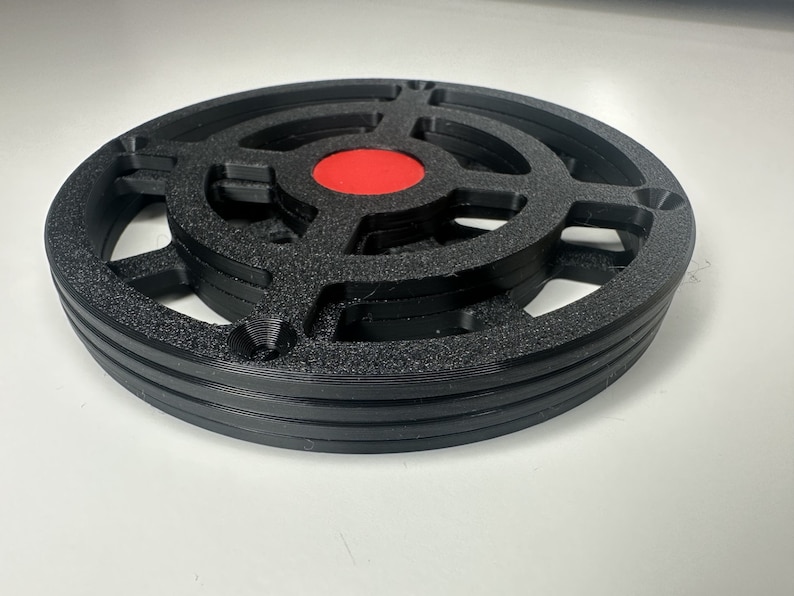 May include: A circular black 3D printed object with a textured surface, featuring a cross-shaped internal structure and a solid red circular center. The object has a layered design with visible horizontal ridges along the outer edge and four holes.