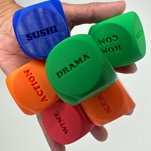 May include: Five colourful dice with different words printed on each side. The words are "Sushi", "Drama", "Action", "Wine", and "Rom Com".