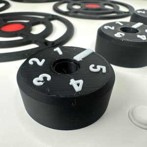May include: Black cylindrical control knobs with white raised numbers one through five and a white indicator line. The knobs are positioned on a white surface alongside black circular grate-like components featuring a central red circular accent.