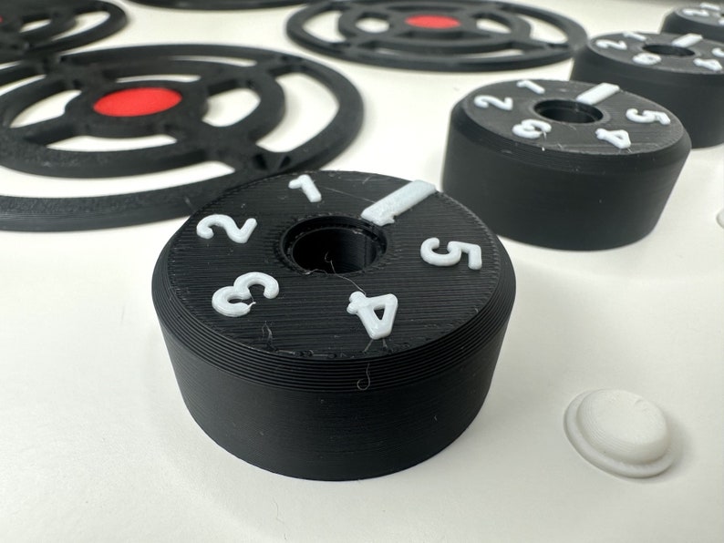 May include: Black cylindrical control knobs with white raised numbers one through five and a white indicator line. The knobs are positioned on a white surface alongside black circular grate-like components featuring a central red circular accent.