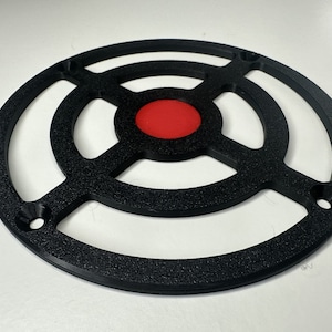 May include: A circular black plastic grill cover featuring a textured finish, a central solid red circular insert, and four evenly spaced mounting holes along the outer ring frame, designed with a cross-shaped support structure.
