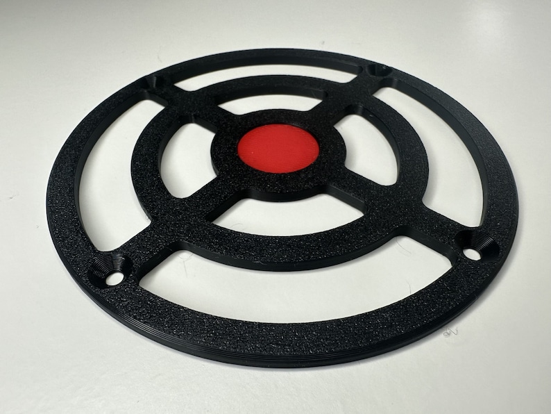 May include: A circular black plastic grill cover featuring a textured finish, a central solid red circular insert, and four evenly spaced mounting holes along the outer ring frame, designed with a cross-shaped support structure.