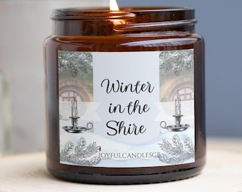 May include: A brown glass candle jar with a white label that reads "Winter in the Shire". The label features a watercolor illustration of a cozy cottage with snow-covered trees and a lit candle.