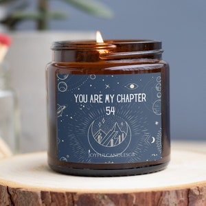 May include: A brown glass candle jar with a blue label that says "You are my chapter 54" with a moon and mountain design. The candle is lit and the flame is visible.