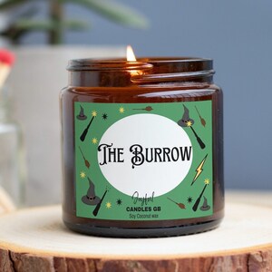 May include: A brown glass candle jar with a green label that says "The Burrow" and features a whimsical illustration of a witch's hat, wands, and stars. The candle is made by Joyful Candles GB and is soy coconut wax.