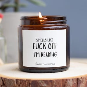 May include: A lit candle in a brown glass jar with a white label. The label reads "Smells Like Fuck Off I'm Reading." The candle sits on a wooden slice. The brand name "JoyfulCandlesGB" is at the bottom of the label.