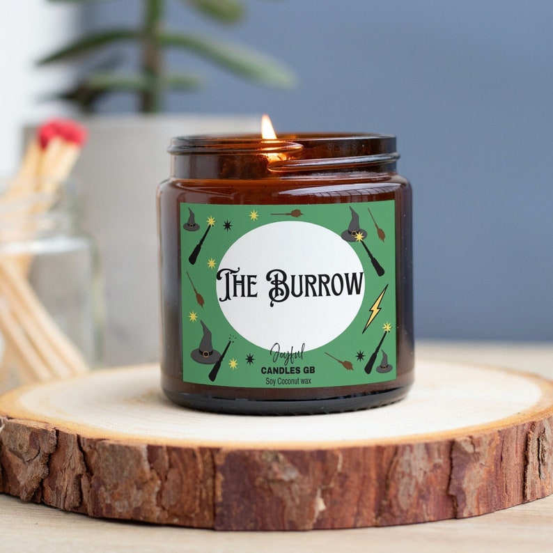May include: A brown glass candle jar with a green label that says "The Burrow" and features a whimsical illustration of a witch's hat, wands, and stars. The candle is made by Joyful Candles GB and is soy coconut wax.