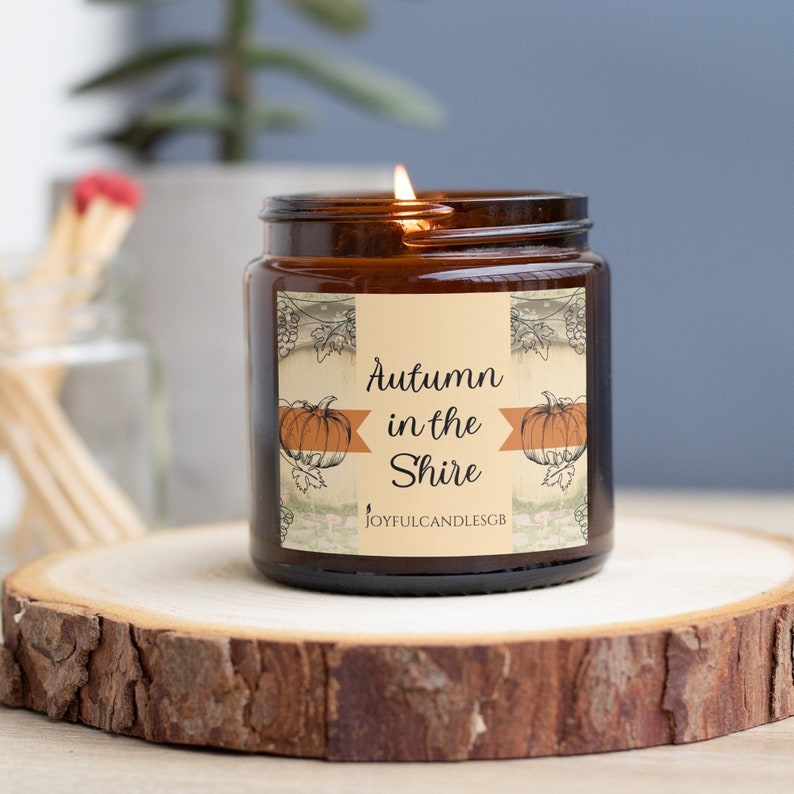 May include: A lit candle in a brown glass jar with a label featuring two orange pumpkins, vine illustrations, and the text "Autumn in the Shire" along with "JOYFULCANDLESGB" printed in black on a cream-colored background.