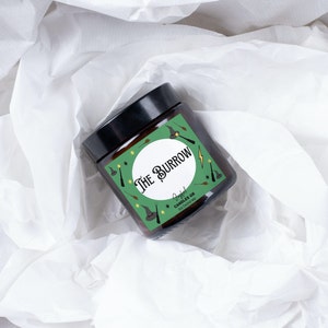 May include: A green and brown candle jar with a white circle and the text "The Burrow" in black. The jar is decorated with a green and yellow pattern of stars, wands, and wizard hats. The text "Soy Coconut Wax" is printed on the label.