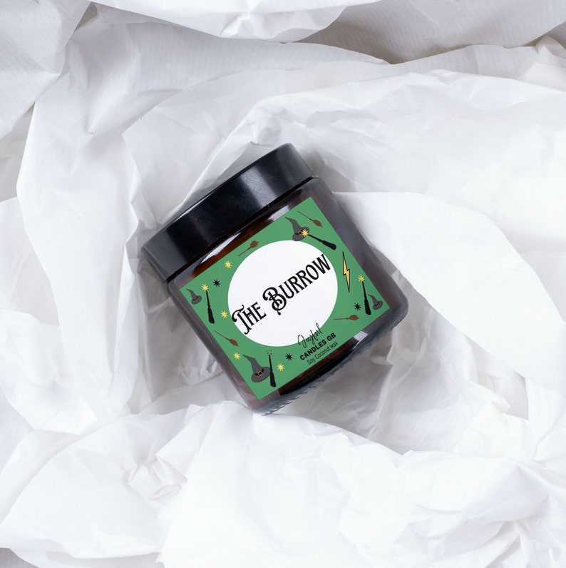 May include: A green and brown candle jar with a white circle and the text "The Burrow" in black. The jar is decorated with a green and yellow pattern of stars, wands, and wizard hats. The text "Soy Coconut Wax" is printed on the label.