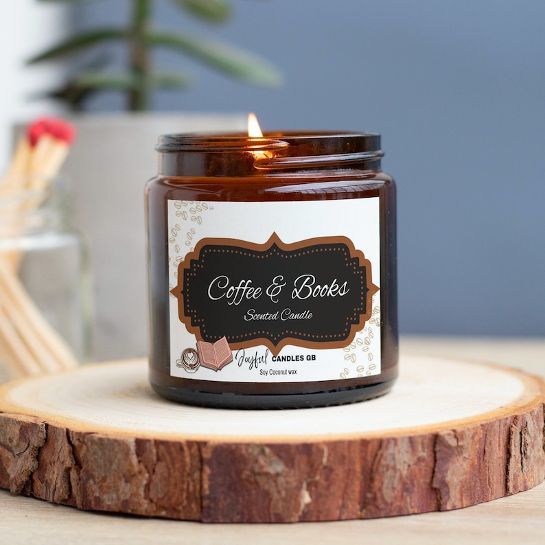 May include: A lit scented candle in a brown glass jar with a label that reads "Coffee & Books." The candle sits on a wooden slice. A small glass jar with matches is in the background.