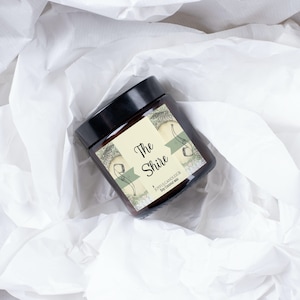 Peut inclure: Un bocal en verre brun pour bougie avec un couvercle noir. L'&eacute;tiquette est cr&egrave;me avec des accents verts et bruns et porte l'inscription "The Shire" avec le texte "Joyful Candles Co. Soy Coconut Wax".