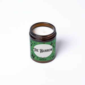 May include: A green and brown candle jar with a white candle inside. The jar has a green label with the text "The Burrow" and "Joyful Candles GB" in white. The label also has a pattern of stars, wands, and witches' hats.