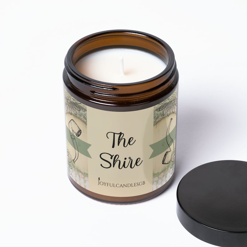 Peut inclure: Un bocal en verre brun contenant une bougie blanche. Le bocal est &eacute;tiquet&eacute; "The Shire" avec un ruban vert et un dessin d'une pipe et d'un seau. L'&eacute;tiquette indique &eacute;galement "JoyfulCandlesGB".