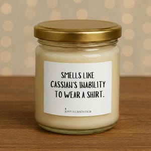 May include: A glass jar candle with a gold lid. The label reads "Smells like Cassian's inability to wear a shirt." The candle is filled with a creamy wax. The brand name "JoyfulCandlesGB" is at the bottom.